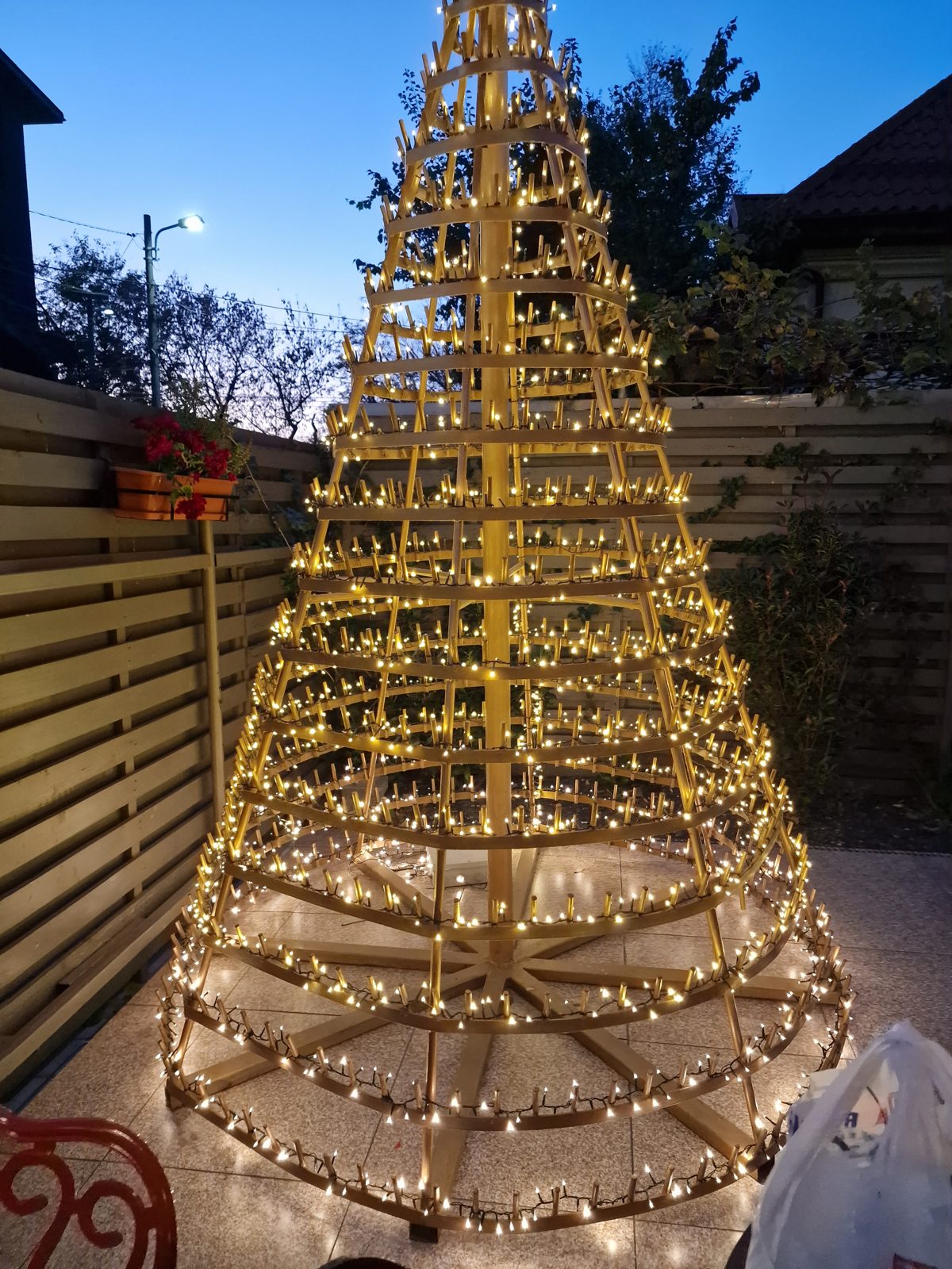 A 10 ft welded steel bottle tree painted gold, wrapped in warm lights, standing on a patio at dusk